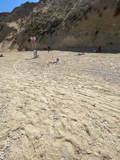 This is a wide-angle, eye-level shot of a sandy beach in Ga'ash, Israel. The beach is backed by a steep, eroded cliff face covered in sparse vegetation. The sand is a light beige color, showing tracks from vehicles and footprints. Numerous small pebbles and shells are scattered across the surface. In the foreground, the sand is textured with tire tracks.
Several people are visible on the beach. In the middle ground, a person lies on their back on a blue towel, sunbathing. Further back and to the right, two people are seated in the shade of the cliff, one reading and the other in a foldable chair. To the left of the cliff, there is a red and white sign with Hebrew text and a symbol depicting a crossed-out wave, indicating no swimming. Two bicycles and a white bag are leaning against the sign. A person is standing near the bicycles, and a small dog is nearby. Further left, another person is lying on a towel. The sky is a clear, bright blue.