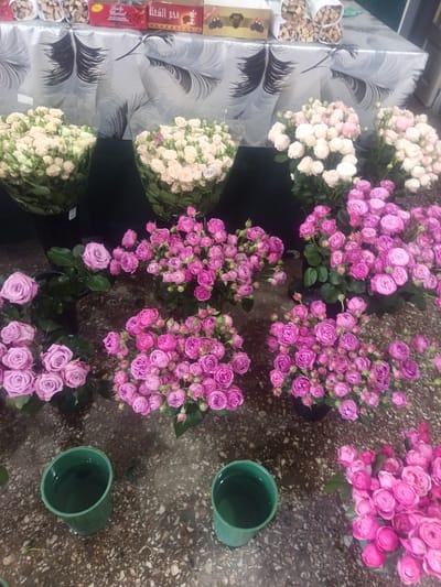 The image shows a display of flowers, primarily roses, in what appears to be a market stall or shop. There are several bouquets of roses in various shades of pink and white. In the foreground, two green cups filled with water are visible on a speckled floor. In the background, the scene suggests a table covered with a patterned tablecloth, with some boxes and bundles of what look like food items. The lighting is somewhat dim, creating a slightly shadowed atmosphere. The overall impression is of a floral vendor's display. There are no people visible in the image. The location is stated as Nor Hachin, Armenia.