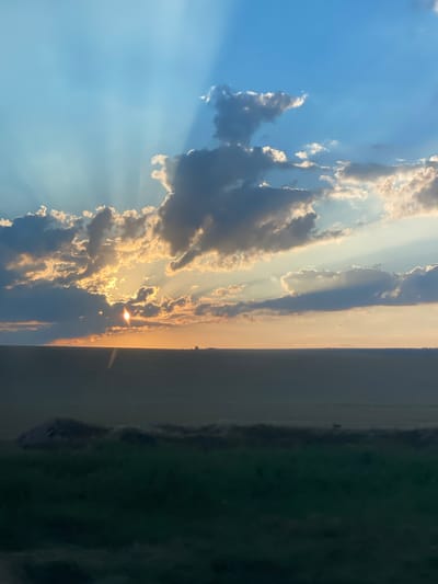 The image captures a sunset over a flat, agricultural landscape. The sun is partially obscured by clouds, casting dramatic rays of light across the sky. The sky transitions from a bright blue at the top to a warm orange and yellow hue near the horizon. Dark, textured clouds are scattered throughout the upper portion of the image, contrasting with the lighter sky. The horizon line is sharp, dividing the sky from the land. The land below appears to be a vast, dark field, with some subtle variations in texture and color suggesting terrain or vegetation. In the foreground, a darker strip of green is visible, possibly a road or a denser patch of vegetation. The overall scene evokes a sense of natural beauty and the end of the day. The image does not contain any identifiable elements that would suggest it is specifically located in the city of Svetlîi, Moldova.