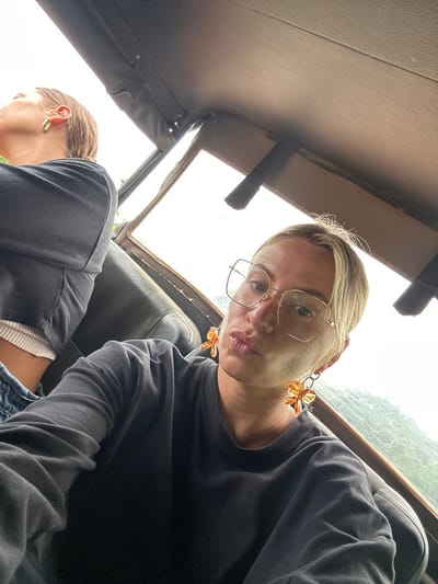 Two women in a vehicle are captured in a low-angle selfie. The woman in the foreground has blonde hair, glasses, and flower-shaped earrings, puckering her lips toward the camera. The other woman, with her head down, wears a grey top and has short brown hair. The vehicle's interior, including the roof, window, and seat, is visible. The background shows a blurred outdoor scene with overcast lighting.