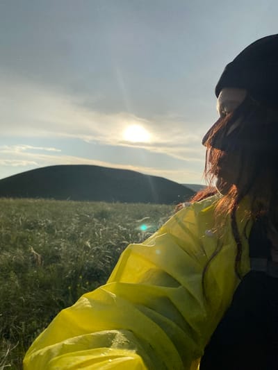 The image shows a person in a bright yellow jacket and black beanie, seen from the side and back, looking out at a natural landscape. The person's face is partially obscured by their hair and the angle of the shot, but they appear to be looking towards the right of the frame. The sun is visible in the sky, partially obscured by clouds and casting a warm glow. In the background, there is a dark, rounded hill or mountain, and a field of tall, wispy grass. The scene appears to be outdoors, possibly in a rural or natural setting. The location is specified as the city of Donskoye, Russia, suggesting this is likely a natural area within or near the city. The lighting and atmosphere suggest it might be late afternoon or early evening.