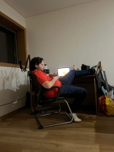 An indoor scene in Pedrouços, Portugal, depicts a male subject seated in a black office chair within a room featuring light-colored walls and a light brown wooden floor. The subject, a man with dark, wavy hair and a beard, wears a red t-shirt and dark blue jeans. He is holding a smartphone in both hands, looking down at its screen. His left foot, clad in a white sneaker with dark stripes, rests on the floor. His right foot, in a similar sneaker, is propped on the top right corner of a light wooden desk.
On the desk, an open silver laptop displays an illuminated, colorful screen. To the left of the laptop are two black audio speakers and a black microphone mounted on a boom arm, positioned towards the left. To the right of the laptop, a clear plastic bottle containing a red liquid is visible, alongside a dark, rectangular object topped with a small metallic ornament. Behind the right side of the desk, a light blue electric guitar with a white pickguard leans against the wall, next to a yellow and red patterned bag on the floor. A window with a dark pane and a wooden frame is visible on the far left wall. Black cables are present on the floor and against the wall near the desk. The room is dimly lit, with light primarily emanating from the laptop screen.