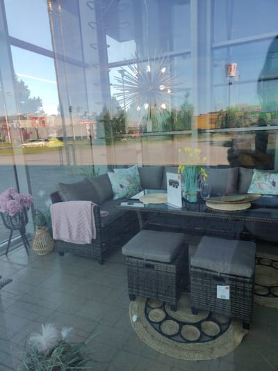 This image depicts a reflection of an outdoor scene through the glass window of a furniture store, likely located in Bielany Wrocławskie, Poland, as indicated by the reflected signage. The foreground is dominated by an outdoor furniture set, including a sectional sofa with dark grey cushions and decorative pillows featuring a tropical bird and leaf motif. A pink throw blanket is draped over one section of the sofa. In front of the sofa is a dark-colored, wicker-style coffee table with a glass top, holding a vase of yellow artificial flowers, a clear glass vase, a brochure or catalog, and decorative placemats. Two matching wicker-style ottomans with grey cushions are positioned in front of the coffee table. The floor inside the store is tiled.
The reflection reveals an urban environment outside. Visible elements include a bright blue sky with a faint contrail, trees, streetlights, traffic lights, and various buildings, including what appears to be a commercial building with large red and white signage, possibly a fast-food establishment or grocery store. A yellow truck with Polish lettering is also visible in the reflection. The overall scene suggests a clear day with ample sunlight, creating reflections on the glass that overlay the interior display. The presence of a chandelier-like light fixture with starburst-shaped elements is also reflected from inside the store.
