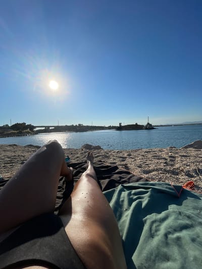 An overhead, wide-angle shot shows a person lying on a pebbled beach next to the clear blue water of Nea Potidea, Greece. The sun shines brightly in the upper left corner of the frame, casting a strong glare on the water. The person's legs are in the foreground, with one leg bent and the other extended. A dark towel and a green towel are spread out on the beach beneath the person. In the distance, a bridge spans across the water, and a lighthouse stands on a rocky outcrop. The sky is a clear, vibrant blue.