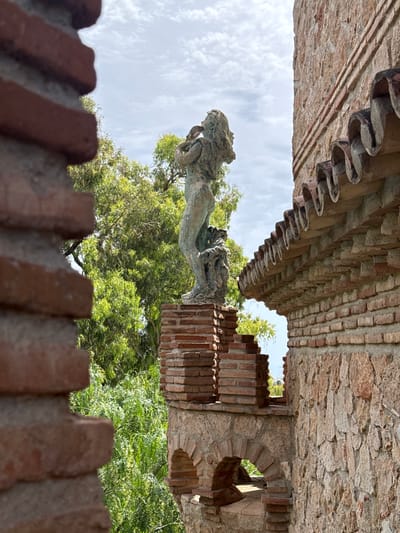 The image shows a bronze sculpture of a person in a garden setting, located in Benalmádena, Spain. The sculpture depicts a figure with flowing hair, holding an object to their mouth as if playing a wind instrument. They are standing on a pedestal made of stacked bricks, which is part of a larger architectural structure. To the right of the sculpture, there is a wall constructed of large, rough-hewn stones with a decorative border of terracotta roof tiles. In the foreground, out of focus, are sections of a brick wall, framing the view of the sculpture and the building. Lush green foliage from trees is visible behind the sculpture and peeking through openings in the stonework. The sky above is partly cloudy. The overall scene suggests a garden or park area within a historic or decorative complex in Benalmádena.
