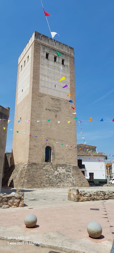 The image depicts a brick tower decorated with colorful triangular flags. The tower features a mix of brick and cement construction, with several windows visible. The sky is clear and blue. The surroundings include other buildings with white walls and tiled roofs. In the foreground, there is a stone path with spherical stone bollards. The image appears to be taken at ground level, looking upwards at the tower. The image's timestamp indicates it was taken on June 1, 2026. The city of Canals, Spain, is identified as the location.