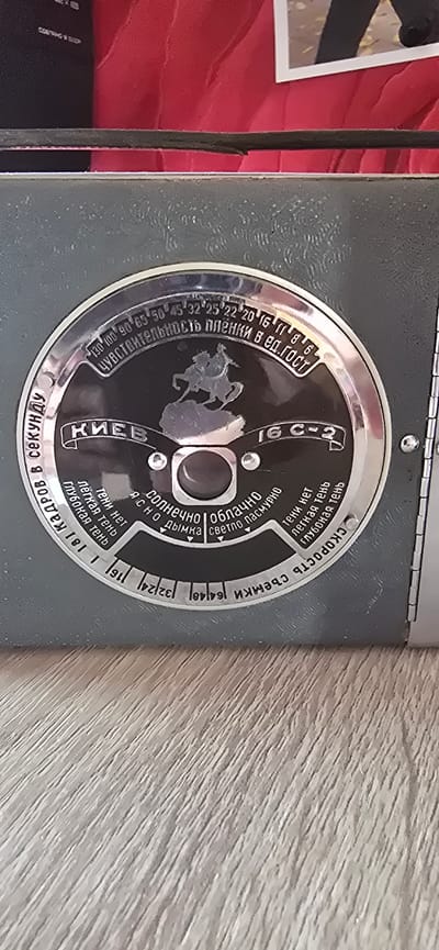 The image is a close-up, high-angle shot of a vintage camera's light meter dial. The dial is circular with a metallic silver rim. The central part of the dial is black and features a raised silver silhouette of a horseman. Text and numbers in Russian are inscribed around the dial. At the top, in a curved arc, is the text "ЧУВСТВИТЕЛЬНОСТЬ ПЛЕНКИ В ЕД. ГОСТ", which translates to "FILM SENSITIVITY IN GOST UNITS," with numbers ranging from 13 to 130. Below this, in a larger font, is "КИЕВ" (KIEV) and "16 С-2". Around the lower part of the dial, other inscriptions include "СКОРОСТЬ СЪЕМКИ" (SHOOTING SPEED), with numbers like 64, 125, 250, and also what appears to be aperture settings like 1.8, 2.8, 4, 5.6, 8, 11, 16. There are also descriptions of lighting conditions such as "СОЛНЕЧНО" (SUNNY), "ОБЛАЧНО" (CLOUDY), "ЯСНО" (CLEAR), "ДЫМКА" (HAZE), "СВЕТЛО ПАСМУРНО" (LIGHTLY OVERCAST), and descriptions of shadows like "ТЕни нет" (NO SHADOWS), "легкая тень" (LIGHT SHADOW), "глубокая тень" (DEEP SHADOW). The dial is set against the dark gray, textured body of the camera. The background is out of focus and appears to be a red, textured surface, possibly fabric, with a sliver of a white-framed picture visible at the very top. The camera is resting on a surface that appears to be made of wood with a textured grain pattern. The image is well-lit, highlighting the details of the camera's dial. There is no indication of the city of Gostagayevskaya, Russia, in the image.