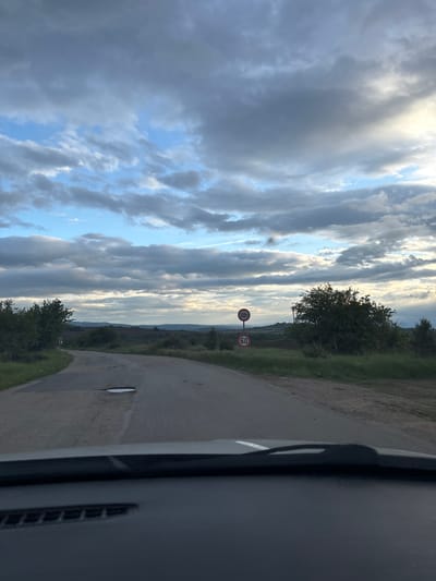 The image depicts a view from inside a vehicle, likely a car, on a rural road in the vicinity of Baylovo, Bulgaria. The road curves to the right. A speed limit sign indicating "60" and another sign stating "20t" (likely a weight restriction) are visible. The sky is filled with a mix of clouds and patches of blue. Vegetation, including trees and grass, lines the side of the road. The car's dashboard is visible in the foreground.