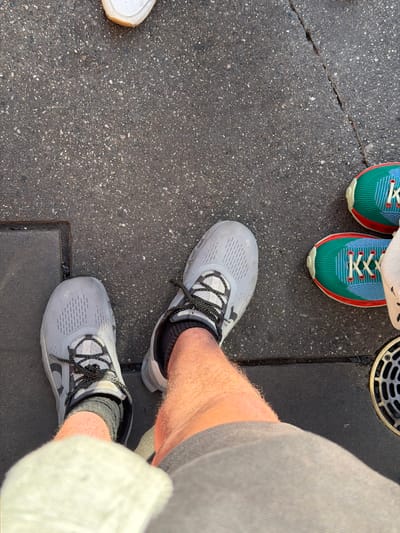 This image, taken in an outdoor setting likely within Anaheim, United States of America, focuses primarily on footwear. The composition is a close-up, high-angle shot of pavement with three distinct pairs of shoes visible.
The nearest pair of shoes are light gray running shoes with black and white patterned laces, worn by someone whose hairy legs are partially visible. The individual is also wearing gray shorts and a white cloth, possibly a towel or bag, is hanging down the side.
A second pair of shoes, positioned further away, are brightly colored running shoes featuring a combination of blue, green, and red hues with white laces. A third, single, white sneaker is partially visible in the lower left corner of the frame.
The ground appears to be a gray, asphalt-like surface with a scattering of small white specks. A metal grate is seen near the top right, adjacent to the colored sneakers, suggesting the location could be a street or sidewalk in an urban area. Overall, the lighting suggests it is daytime.