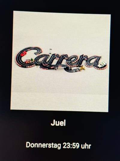 This image displays a photograph of an electronic screen, visible by the distinct pixel grid. The screen presents a digital image of a miniature slot car racing track arranged to spell the word "Carrera." The track segments are predominantly black, edged with red and white stripes. A red slot car is positioned on the initial "C," while a white slot car rests on the final "a." Embedded within the track layout are miniature landscape elements, including a small, red-roofed building and green trees. The background of this displayed image is a uniform light beige or off-white. Below the displayed image, on the screen's dark background, the text "Juel" is visible, followed by "Donnerstag 23:59 uhr," indicating the scene is observed in Vellmar, Germany.