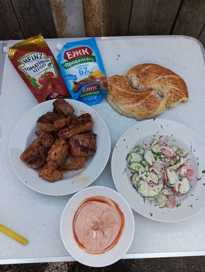 The image shows a table set outdoors with various food items. There's a plate of grilled meat, a plate with a salad of cucumbers, tomatoes, onions and dill, a bowl of mixed ketchup and mayonnaise, a flatbread, and containers of Heinz tomato ketchup and EЖK mayonnaise. The background includes a wooden fence and a metal post. The setting appears to be a casual outdoor meal, likely in a backyard or picnic area. While the food items are Russian brands, the city of Кодинка cannot be determined from the image.