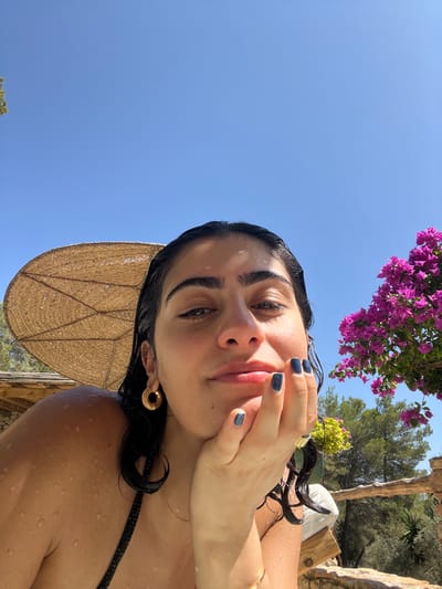 The media file is a self-portrait of a young woman taken outdoors on a sunny day. The woman has dark hair, which appears wet and is styled to frame her face. She is wearing a black bikini top and has distinctive large, gold hoop earrings. Her fingernails are painted a dark navy blue, and she is touching her face with her right hand. Water droplets are visible on her skin and hair, suggesting she may have recently been swimming or showering.
Behind her, a large straw umbrella is partially visible, adding to the impression of an outdoor leisure setting. The background includes lush green trees and a bright blue sky, with a cluster of vibrant pink bougainvillea flowers hanging from above and to the left of the frame. The lighting and clear sky suggest it is daytime, likely late morning or afternoon. The overall mood appears relaxed and summery. The image was taken in Santa Eulària des Riu, Spain. There is no visible text in the image.
