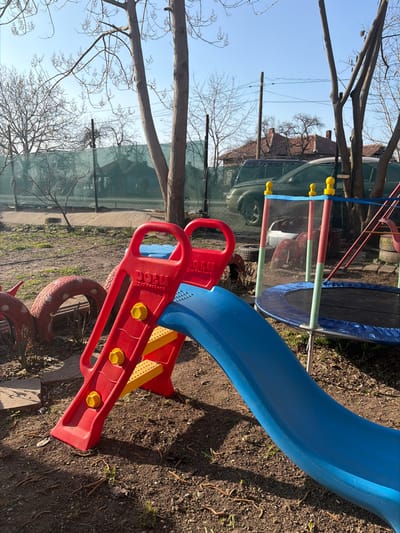 This image captures an outdoor play area located in Kriva Bara, Bulgaria, under a clear blue sky. In the foreground, a red and yellow plastic children's slide, branded "Dolu Toy Factory," is positioned on loose brown soil. Adjacent to the slide on the right, a circular trampoline with a blue spring pad and a black jumping surface is present, enclosed by a blue safety net supported by poles topped with yellow caps. To the left of the slide, several red-painted tires are partially visible on the ground, arranged in a curving pattern. The ground cover consists primarily of bare soil with scattered small green sprouts and dry organic debris. Numerous deciduous trees, currently devoid of leaves, are distributed across the scene, with their shadows cast by direct sunlight. In the background, beyond a green mesh fence, residential structures with tiled roofs and at least two dark-colored sport utility vehicles (SUVs) are visible. Utility poles and overhead wires are also present against the sky. No individuals are depicted in the scene.
