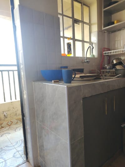 The image depicts a kitchen area in Limuru Central ward, Kenya. A light-colored tiled countertop is visible, with a blue bowl and a blue patterned mug on it. Behind the countertop, a kitchen sink with a faucet is situated below a window with multiple panes. A dish rack with clean dishes is to the right of the sink. Above the sink area, a shelf holds a few items, including what appears to be a yellow tin. To the left of the kitchen counter, an open doorway reveals a tiled floor and a metal railing outside. The overall scene suggests a functional, though somewhat cluttered, kitchen environment.