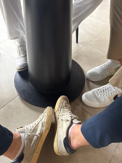 The photo shows the feet of three people around a black table base. Two people wear white sneakers; one is a newer style, the other appears worn. The third person sports dirty white sneakers, with navy blue heels and beige soles, and is wearing short socks. The floor is tiled and off-white. The lighting is bright.