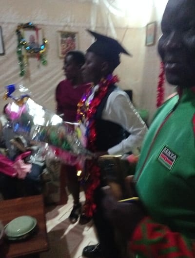 Inside a decorated room, three people are visible. A graduate wears a cap, a white shirt, a vest, and a red and silver garland. A woman stands nearby in a burgundy top. In the foreground, a person wears a green jacket with a Kenya flag patch. Decorations adorn the walls, including tinsel and framed pictures. A wooden table with a plate sits on the lower left. The image suggests a celebration or graduation event.