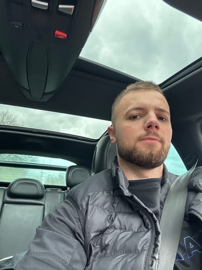 A male individual is depicted seated in the front of a vehicle. The scene is situated in the city of Брусовeте, Bulgaria. The subject, appearing to be in his 20s or early 30s, possesses short, light brown hair and a trimmed light brown beard. He wears a black puffer jacket over a dark crew-neck shirt and is secured by a grey seatbelt across his chest and lap. His expression is neutral, and he looks directly towards the camera. The vehicle's interior is predominantly dark, featuring black leather or synthetic seating, a black headliner, and an overhead console equipped with a visible red "SOS" button. A panoramic sunroof above reveals an overcast, grey sky. Through the rear window, a generic outdoor environment is discernible, consisting of bare trees, distant vegetation, and faint power lines or utility structures, providing no specific identifying landmarks for the precise location within Брусовeте.