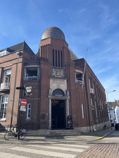 This image captures the architectural detail of the former Post Office building in Maastricht, Netherlands, situated on an urban street. The structure is predominantly constructed of red brick, with a prominent rounded tower. A sign above the entrance reads "Post Telegraaf". There is an ornate emblem positioned above the entrance, and an awning over the doorway, through which there is a glimpse of an elaborate chandelier inside.

In the foreground, there is street signage indicating traffic regulations, along with street name signs, and parked bicycles. The pavement in the foreground has a crosswalk pattern. The sky above is clear and blue.