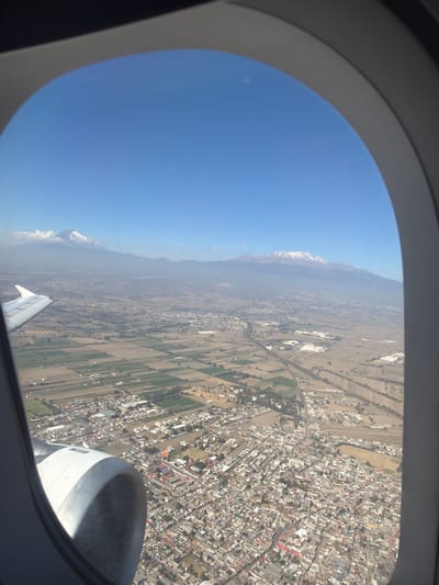 An aerial view from an aircraft window captures a clear daytime scene above Santa Ana Xalmimilulco, Mexico. The gray aircraft window frame partially encloses the image. In the lower-left foreground, a section of a white aircraft wing is visible, extending towards the left, with an adjacent white jet engine nacelle featuring a dark intake opening. Below the aircraft, the city of Santa Ana Xalmimilulco is spread out, characterized by a dense grid of numerous residential and commercial buildings with predominantly light-colored roofs, connected by roads. Adjacent to and within the urban development are segmented agricultural fields, presenting a mosaic of green and brown plots. In the background, under a clear blue sky, a range of mountains is discernible through a light atmospheric haze. Two prominent snow-capped peaks, consistent with Popocatépetl (left, with some cloud cover) and Iztaccíhuatl (right), rise above the distant landscape. No individuals are visible.