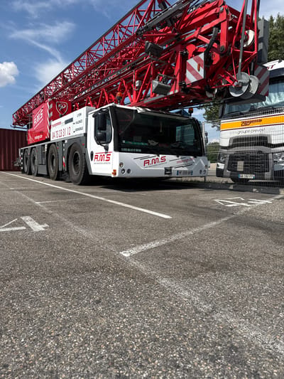 This media file shows a large red crane truck parked on an asphalt surface, likely a parking lot or industrial area. The truck is white with red accents and features the logo "R.M.S" prominently displayed on its side, along with text indicating "LEVAGE MANUTENTION CONSTRUCTION" and phone numbers. It also has the website "rmslevage.com" visible. The truck's license plate is "HF-050-AH."
Behind the crane truck, another vehicle with a yellow and white striped trailer is partially visible, bearing the website "www.groupe-prevost.com" and the license plate "GA-182-TP." The crane itself is a massive, intricate red metal structure, suggesting it's a piece of heavy machinery used for construction or industrial lifting.
The scene appears to be outdoors on a clear, sunny day, as indicated by the bright light and the blue sky with scattered clouds. The asphalt surface has white painted lines, possibly demarcating parking spaces or lanes. The location is identified as Grenay, France. There are no people visible in the frame. The overall impression is one of a static industrial setting, with the focus on the large equipment.