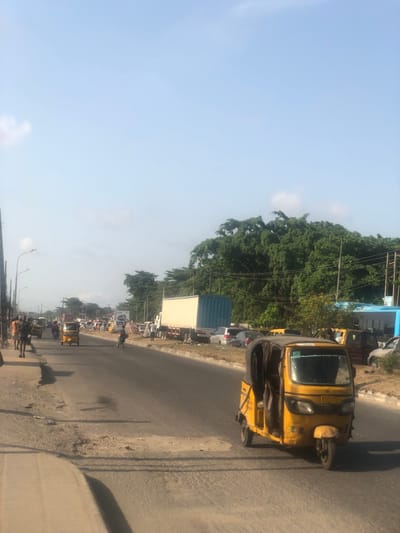 The image depicts a daytime street scene in Alasia, Nigeria, under a clear sky. In the immediate right foreground, a yellow motorized trishaw (keke napep) with a black fabric cover is positioned, facing towards the left. The paved, two-lane road displays visible wear, including potholes and asphalt repairs. Further down the road, multiple vehicles, including other yellow trishaws, motorcycles, and cars, are present. A large white semi-trailer truck with a blue shipping container is parked along the right shoulder. Several pedestrians are observed walking or standing on the left sidewalk and roadside. The right side of the street features a collection of parked cars, some appearing to be in disrepair, and a light blue building structure. Utility poles with streetlights line the left side of the road. Dense green foliage forms the horizon in the background beneath a mostly clear, light blue sky with minimal cloud cover.