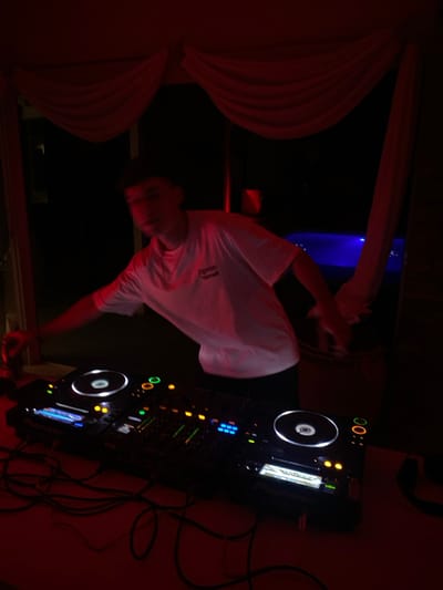 A DJ stands at a DJ controller, wearing a white t-shirt in a dimly lit room. The room is accented with red and blue lights. A large, dark object looms in the background, partially obscured by a white curtain. A tangle of cables and cords runs alongside the controller, suggesting a complex sound setup. The composition is vertically oriented.