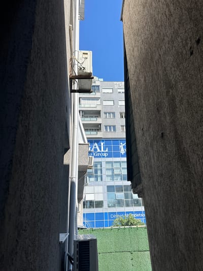 The image shows a view upwards from a constricted space between buildings, likely an alley or narrow courtyard. The composition is dominated by the concrete structures above and below the frame, suggesting an enclosed area. Visible between the buildings are sections of two distinct buildings: one is a modern, glass-facaded building with blue and white signage (partially obscured but shows "Te Group" and "Dëmet Shëndetsp"), and the other is a grey building with symmetrical window patterns. On the top of the lower building are various utility objects, pipes, and air conditioning units. The sky is a clear blue, visible through the opening in the buildings. There are no people visible. The image appears to be taken in Pristina, Kosovo, based on the business names on the building.