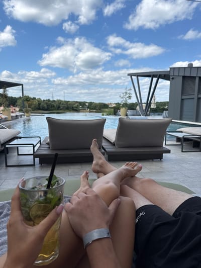 An outdoor scene captured from a first-person perspective depicts two individuals lounging poolside at a resort in Oseshchyna, Ukraine, under a blue sky with scattered cumulus clouds. In the immediate foreground, a person's right hand, with light-colored manicured fingernails, holds a clear glass containing a green beverage with lime slices, mint leaves, and ice, served with a dark straw. The lower legs and feet of two individuals are visible, indicating they are reclining on lounge furniture. The left pair of legs, appearing female, has an ankle bracelet on the right ankle and light pink nail polish on the toes. The right pair of legs, appearing male, wears black shorts with a partially visible "Gymb" logo and a grey wristband with the number "7" on the right wrist. The individuals' hands are resting on or near each other's thighs.

The foreground is a light grey tiled patio area where a grey towel is draped over a lounge cushion. To the left, a small black-framed side table with a white top is visible. Beyond the reclining individuals, a large, blue infinity-edge swimming pool dominates the midground, with several unoccupied dark-framed lounge chairs featuring grey cushions arranged around its perimeter. On the right side of the pool, modern dark-colored buildings with distinct angled support structures and horizontal slats are visible. In the background, a large natural body of water, consistent with a lake, extends across the horizon, bordered by a dense line of trees and distant structures, including a utility pole.