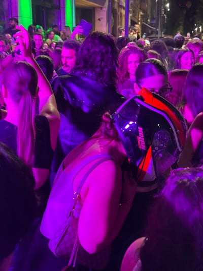 A crowd gathers in a dimly lit space with purple and green lights. A woman in a white top has a black Nolan motorcycle helmet on her head, partially obscuring her face. Some individuals are holding up phones, and a woman in a black leather jacket stands nearby. The crowd appears to be at a concert or event.