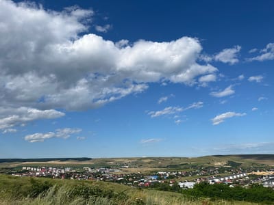 A wide, outdoor, horizontal shot captures a clear blue sky with scattered white clouds above a hilly landscape. In the mid-ground, a dense town or village, identified as being in Piatra-Neamț, Romania, is visible. The buildings are a mix of multi-story apartment complexes with white facades and red roofs, and smaller residential houses with red, brown, and white roofs. The town is nestled within rolling green and yellow hills, with some areas appearing to be agricultural fields. The foreground features a grassy slope with tall, dry grass and some low-lying green vegetation, suggesting a vantage point from a hill overlooking the town. The overall impression is a sunny day in a suburban or rural setting.