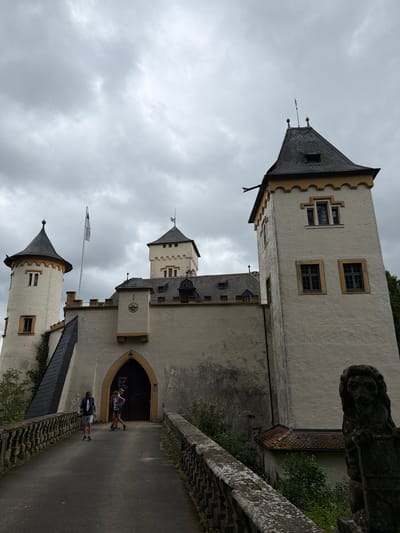 A castle stands on a hill against a cloudy sky. The castle is white with a yellow trim and dark grey roofs. It has several towers, including one on the right with a tall, pointed roof and two smaller towers on the left. A drawbridge leads to the main entrance, which is an arched doorway. Two people are walking across the drawbridge, one in a blue shirt and shorts, and another in a red shirt and dark shorts. A third person in a grey vest and shorts is walking away from the camera on the drawbridge. A stone lion statue stands to the right of the drawbridge. The castle appears to be well-maintained and is surrounded by trees and greenery. The overall impression is one of history and grandeur.