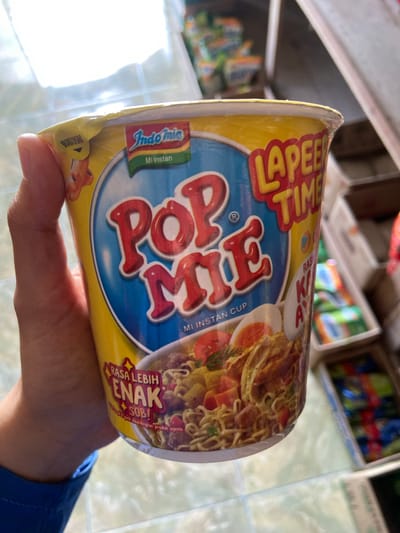 A person's hand is holding a yellow cup of instant noodles. The cup is branded "POP MIE" with "Indomie" logo and "Mi Instan" printed on it. The noodles are shown in the picture with toppings such as boiled egg and vegetables. The background shows shelves filled with other products, suggesting the image was taken in a store or market. The lighting suggests an indoor environment, possibly a shop in Sekar Mawar, Indonesia, given the branding of the product. The cup appears to be sealed with plastic wrap.
