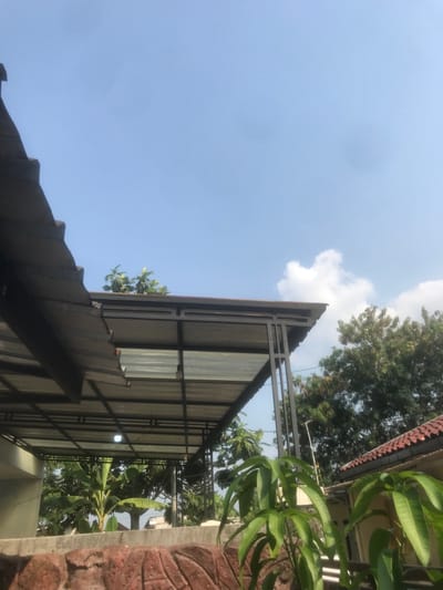 The image displays an outdoor scene in Limbangan Sari, Indonesia, characterized by clear blue skies with scattered white cumulus clouds. The foreground features a reddish-brown textured wall. Directly behind this wall, on the right, two plants with prominent green leaves are visible. A light-colored building wall is partially visible on the far left.
A dark-framed metal canopy with a translucent corrugated roof extends across the mid-ground, supported by vertical metal pillars. A single white light fixture is suspended beneath the canopy structure. Above this canopy, several small green leafy plants are visible. To the upper left, a portion of a dark corrugated metal roof is present.
In the background, to the right of the canopy, a cluster of mature green trees is visible. To the far right, a section of a residential building with a reddish-brown tiled roof is partially visible, with a thin metallic antenna or pole positioned in front of it. Further back on the left, under the canopy, a large banana plant is identifiable among other green foliage. No individuals, actions, or specific interactions are depicted.
