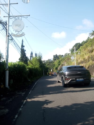 The image depicts a road in an area named Ar-Raafi, Indonesia, with a dark-colored car traveling on it. Road signs are visible on posts along the roadside. Lush green vegetation covers the slopes to the right, and trees line the left side of the road. Other vehicles can be seen in the distance. The sky is blue with some clouds. "B 1636 SDW" is visible on the license plate of the car.