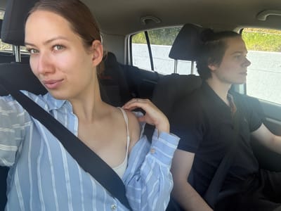 Two individuals are seated in what appears to be a car, seen from an upward angle as if the camera is positioned below them. The image is bright, suggesting daylight.

On the left side of the frame, one person with dark hair, possibly tied back, is visible in profile. They are wearing a dark, possibly black, long-sleeved top and have a seatbelt fastened across their chest. Their gaze is directed forward or slightly downward.

On the right, a second person with brown hair, pulled back, looks directly towards the viewer with green eyes. They are wearing a light blue and white striped long-sleeved shirt with silver, circular buttons on the cuffs. A black seatbelt is visible across their shoulder and chest. Underneath the striped shirt, a white bra or bikini top is visible. Their right hand is shown adjusting this white undergarment, while a silver ring is visible on the ring finger of their left hand. Their expression is neutral.

Through the car windows in the background, bright sunlight illuminates an outdoor scene with green foliage and light-colored structures, indicating clear weather. The car's interior includes dark headrests and a tan-colored ceiling panel with a handle. No visible text is present in the image. The setting is a car interior in Obzovica, Montenegro.