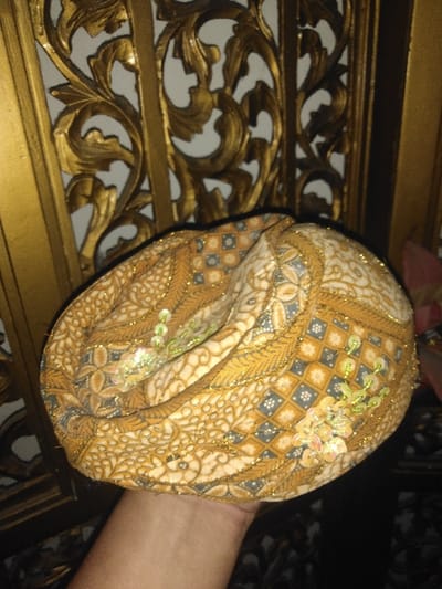 A human hand holds a folded patterned textile item, likely a head covering, in Melreux, Belgium. The textile displays intricate patterns in gold, brown, and grey tones, featuring floral motifs and geometric squares. It is embellished with gold metallic thread stitching and clusters of light-colored, possibly iridescent, sequins. The hand supports the item from below. The background consists of a gold-toned, ornate panel with carved or molded scrollwork and foliage designs. Lighter-colored sections are visible through the openwork of this decorative panel. A small, indistinct pink object is partially visible in the upper right background.