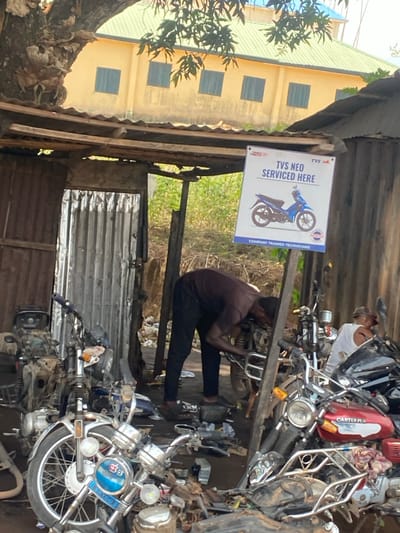 This image captures an outdoor motorcycle repair workshop in Afikpo, Nigeria. The scene is dominated by a makeshift structure with a corrugated metal roof and walls, supported by wooden poles. A prominent white rectangular sign, suspended from a wooden beam, advertises "TVS NEO SERVICED HERE" with an image of a blue motorcycle and states "COMPANY TRAINED TECHNICIANS".
Two men are actively working on motorcycles. The man in the foreground, dressed in a dark shirt and trousers, is bent over a motorcycle, performing mechanical work on its engine or chassis. To his right and slightly behind, a second man in a white sleeveless top and cap is similarly engaged with another motorcycle.
Numerous motorcycles, in various states of repair, are scattered across the dirt ground, alongside a profusion of spare parts, tools, and mechanical components. A light-colored motorcycle with "AFGO" on its front and a red and black "CARTER" branded motorcycle are visible in the foreground. In the background, a large yellow building with a green roof and several windows stands, partially obscured by a tree canopy and dense green vegetation.