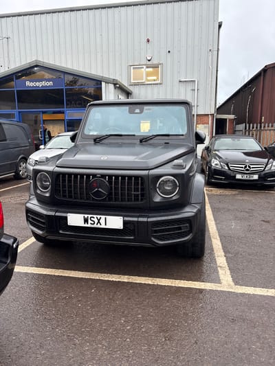 The image depicts an outdoor commercial parking lot at Dach Coach Sales Ltd, located at The Old Brickworks, Nibley, United Kingdom, under an overcast sky. The asphalt ground is wet and delineated by yellow parking bay markings.

In the central foreground, a black Mercedes-Benz G-Wagen SUV is parked, its front facing the viewer. It features a black grille with a black Mercedes-Benz star emblem, which displays a red central element. The front license plate reads "WSX 1". To the immediate left of the G-Wagen, a dark grey Mercedes-Benz Vito van is partially visible. To its right and slightly recessed, a black Mercedes-Benz E-Class sedan is parked, bearing the front license plate "V1 KBH". A small segment of a red vehicle is present on the extreme left edge of the frame.

The background consists of two buildings. The primary structure, to the left, features light grey corrugated metal siding. A glass-fronted entrance section is labeled "Reception". A single individual, clad in a dark garment, is partially discernible inside the reception area. A red security alarm device is mounted on the building's wall, above which a small, internally illuminated window is visible. To the right, a second building, characterized by reddish-brown corrugated metal siding, is partially obscured by a metal fence.