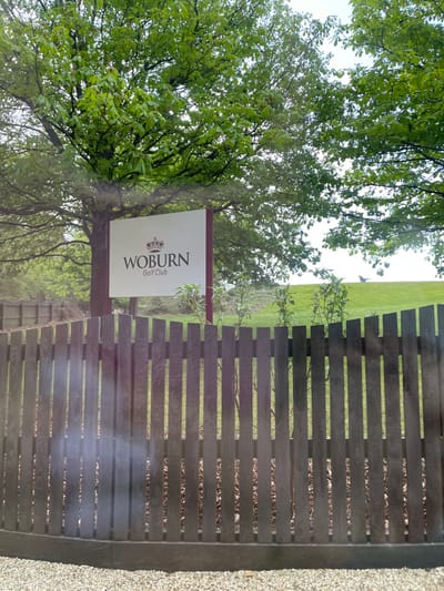 The image shows a wooden fence with a sign attached to it. The sign is white with black lettering and depicts a crown above the word "WOBURN" and below the words "Golf Club." The sign is attached to a maroon post. Behind the fence, there is a grassy area. The fence is made of vertical wooden boards that are closely spaced. There is a small patch of gravel visible at the bottom of the image.  The coordinates provided (latitude: 51.99, longitude: -0.662) place the location in the city of Woburn, Bedfordshire, United Kingdom. There are no people visible in the image.