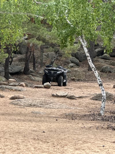 An all-terrain vehicle (ATV) is parked on a dirt path amidst a rocky, wooded area. The ATV is black and appears to be a utility model with a roll cage. It is positioned centrally in the frame, facing slightly to the left. The ground is covered in dry dirt and small stones, with larger rocks scattered throughout. Several trees are visible in the background, some with dark bark and sparse green leaves, and one prominent birch tree with white, mottled bark in the right foreground. The overall impression is a natural, undeveloped landscape. The location is the city of Aiyrtau, Kazakhstan.