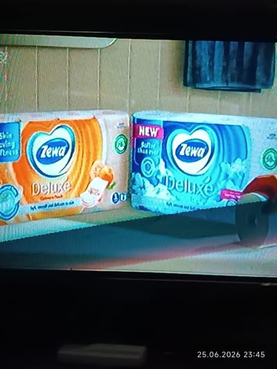 The media file displays two packages of Zewa Deluxe paper towels against a tiled, light-colored wall. A dark-colored towel hangs on the wall to the right. The left package is predominantly orange with blue accents, while the right package is blue with white accents and a pink "NEW" label. Both packages feature a heart-shaped logo with the Zewa brand name. The left package also has text that appears to read "Skin Loving Softness" and "Deluxe". The right package has text that reads "NEW", "Softer than ever", and "Deluxe". The scene is well-lit, suggesting it might be daytime or indoors with good lighting. There are no people visible in the frame. The setting appears to be a bathroom or a kitchen due to the presence of towels and paper products. A timestamp in the lower right corner indicates "25.06.2026 23:45". The image quality is somewhat blurry, typical of a photograph taken from a screen. The overall mood is neutral and commercial, focusing on the product presentation.