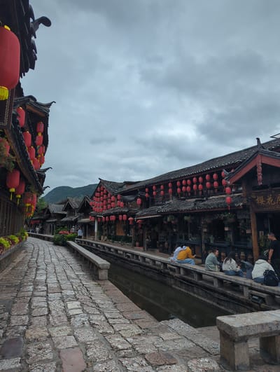 This image captures a scene in the Gucheng District of China, likely an ancient town. The architecture is traditional, with dark tiled roofs and wooden structures. Numerous red lanterns are hung along the buildings and on display, suggesting a festive or cultural event. A cobblestone path leads along a canal, bordered by stone walls.
Several people are visible, some walking along the path or seated on a wooden balcony overlooking the canal. One person is wearing a yellow top, another a white jacket, and others are in darker clothing. Their postures and the casual arrangement of people suggest a relaxed, everyday atmosphere rather than a specific event.
The sky is overcast, indicating a cloudy or perhaps drizzly day. The light is diffused, and there are no strong shadows, suggesting it is daytime, possibly late morning or afternoon. The greenery interspersed with the buildings and along the path adds a touch of nature to the urban landscape.
A sign with Chinese characters is visible on one of the buildings, but the text is not clearly legible. The overall impression is one of a charming, historic location, possibly a tourist destination, characterized by its traditional architecture and abundant red lanterns.