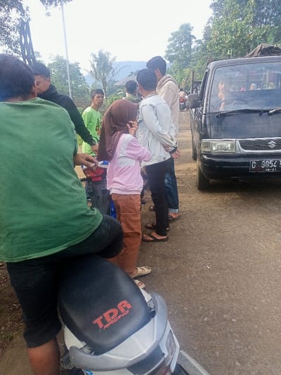 A daytime scene captures a group of people gathered on a rural road in Kecamatan Pangalengan, Indonesia. In the foreground, a person is seated on a white and silver motorbike, facing away from the camera. The motorbike's seat is black with the red text "TDR RACING" visible.
A cluster of individuals stands in the middle ground, mostly young adults. On the left, a person in a green t-shirt looks towards the camera. A young woman, dressed in a brown hijab, a pink long-sleeved shirt, and brown trousers, stands with her back mostly to the viewer, appearing to observe something to her right. Other men in casual attire, some wearing sandals, are also part of the gathering.
To the right, a black Suzuki Carry-type pickup truck is positioned on the road. The driver, a man, is visible through the windshield, looking towards the left. The truck's license plate, "D 9854 H," along with "08-21" (likely indicating August 2021), is clearly visible on the front bumper.
The background features abundant green trees and foliage, with distant hills or mountains under a bright, slightly hazy sky. The road appears to be unpaved or roughly paved, consistent with a rural setting. The scene suggests a casual stop or gathering along the road.