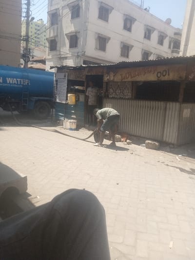 The image captures a daylight street scene in Tudor ward, Kenya. A blue water tanker truck, marked "N WATERF" and displaying a phone number, is parked on a paved surface on the left. A thick black hose extends from the truck across the light-colored interlocking brick pavement. A male individual in a light green t-shirt and dark shorts is bent over, actively handling the hose near a single-story structure constructed from corrugated metal and wood. This structure features the yellow graffiti "LOLLIPOP 001" on its facade. Another male individual, wearing a light-colored, possibly patterned, t-shirt, stands within the doorway of this structure. A yellow jerrycan with a spigot and two white buckets are positioned on the ground outside the structure. In the background, a multi-story white building with numerous windows is visible under clear skies. The lower portion of a person's leg, clad in dark trousers, is present in the immediate foreground, indicating the perspective of an observer or photographer.