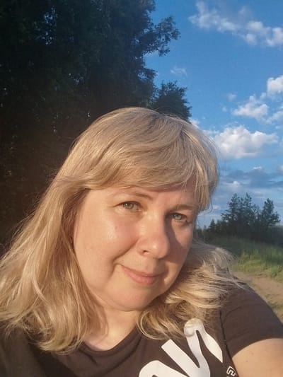 In a close-up selfie taken outdoors, a fair-skinned woman with blonde hair and green eyes smiles at the camera. She is wearing a dark t-shirt with a white graphic on it. Behind her, a landscape of trees and a blue sky with white clouds can be seen, suggesting the photo was taken in a rural or park-like setting. The lighting indicates it is likely late afternoon or early evening. There are no specific landmarks or contextual clues to place this image in Chkalov, Russia.