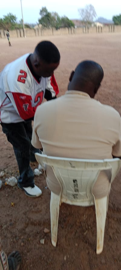 Two men are the focus of the image. One man, wearing an Atlanta Falcons jersey with the number "2" visible, is leaning toward the other, who is sitting in a white plastic chair. The seated man wears a tan shirt. The background shows a dirt field with scattered rocks and a few trees. The scene appears to be outdoors, possibly in a park or recreation area.