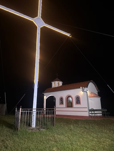The image captures a nighttime scene featuring a large, illuminated cross in the foreground, set against a dark sky. The cross is white and outlined with yellow lights, with wires extending from its arms to anchor points in the ground. To the right of the cross is a small, white chapel with a red-tiled roof. The chapel has arched windows and decorative religious icons above them. In front of the chapel are two dark benches. A metal fence surrounds the base of the cross, and the ground is covered in green grass. The location is identified as being in the city of Parvomai, Bulgaria.