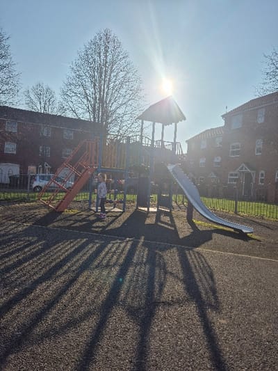 An outdoor playground scene is depicted under bright sunlight, indicating either morning or late afternoon due to the long, distinct shadows cast across the dark grey, granular safety surfacing in the foreground.
**Location:** This residential playground is situated within the city of Hereford, United Kingdom. While the precise street or estate is not visually identifiable, the background architecture is typical of UK housing developments.
**Subjects and Objects:**
* **Playground Structure:** A multi-component structure dominates the mid-ground. It features a central elevated platform with a gabled, reddish-brown roof, accessible via orange open-tread stairs on the left. A grey, straight slide descends from the platform towards the right. Blue metal support poles and railings are visible.
* **Child:** One young child stands on the safety surface beneath the playground structure, near the base of the orange stairs. The child wears a light-colored, patterned top, dark trousers, and pink shoes, and is positioned facing generally towards the right.
* **Buildings:** Two multi-story (at least three visible floors) red brick residential buildings, likely apartment blocks, form the background. They feature numerous white-framed windows and white-framed ground-floor entranceways/porches. The building on the right has a terracotta-tiled roof.
* **Vegetation:** Several tall, leafless deciduous trees are visible in the background, positioned between the playground and the buildings. One large tree stands prominently behind the playground structure.
* **Fencing:** A low, green metal fence separates the playground area from the residential properties.
* **Vehicle:** A white passenger car is partially visible parked behind the fence on the far left.
**Environment and Actions:**
The sky is clear and bright, with the sun positioned high in the upper-center/right, causing significant lens flare and illuminating the scene from behind the playground equipment. The child is static, not actively engaged in play. No other individuals are present. The overall environment suggests a well-maintained urban or suburban play area.