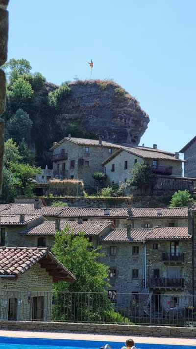 The image shows a view of the town of Rupit i Pruit, Spain, featuring traditional stone houses with red tile roofs nestled into a hillside. A large, weathered rock formation dominates the upper background, topped with a flag. Lush green trees and vegetation surround the buildings and the rock. In the foreground, a bright blue swimming pool occupies the bottom portion of the frame, with a glimpse of two people, one child and one adult, sitting on the edge of the pool. The scene depicts a sunny day with a clear blue sky.