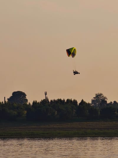 The image captures a powered paraglider in flight against a pale, hazy sky. The paraglider, consisting of a brightly colored wing with yellow, green, and red accents, is positioned in the upper left quadrant of the frame. Below it, a small, dark frame with what appears to be two occupants is suspended. The scene below the sky shows a dark line of trees forming a dense forest, with hints of a body of water in the foreground. The water has a rippled surface reflecting the muted tones of the sky. A tall, slender telecommunications tower is visible amongst the trees on the left side of the frame, and a smaller, darker vertical structure is also present to the left of the tower. The overall impression is one of an outdoor recreational activity taking place over a natural landscape, possibly near a body of water, with man-made structures present in the distance.